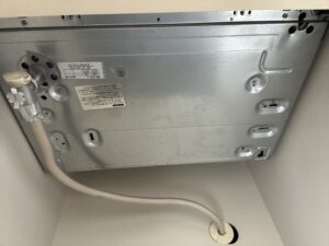 キッチン流し台の下から見上げたビルトインコンロの底部。接続されたフレキ管やガス栓が見える状態で、交換前の確認用に撮影された写真。