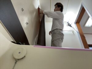 ユニットバスの壁面にダイノックシートを施工中の職人。木目柄シートでビジネスホテル風に仕上げる様子